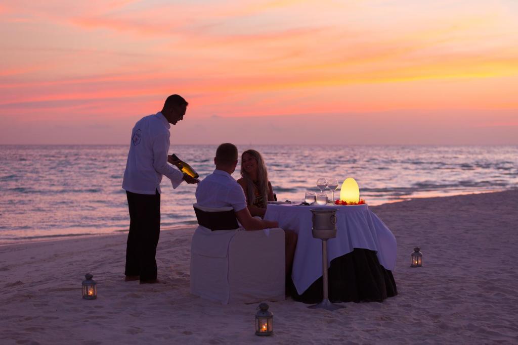 Romantisks vakariņu galds pludmalē ar saulrietu fonā, kur viesmīlis pasniedz šampanieti diviem cilvēkiem. Galds ir dekorēts ar ziediem un svečturiem, radot intīmu atmosfēru.