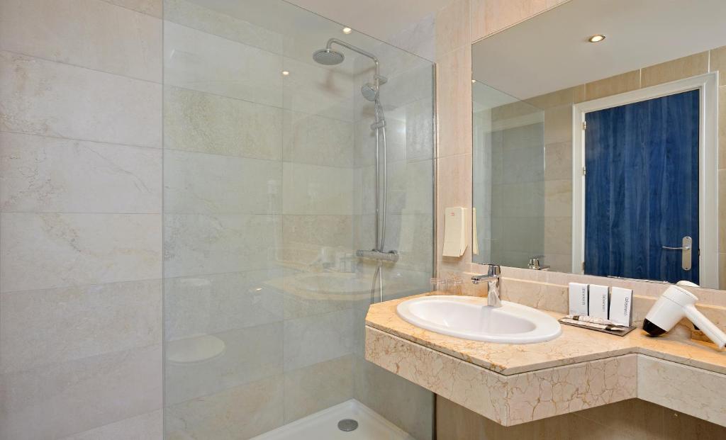 Mūsdienīgs vannasistabas interjers ar stikla dušu un elegantu izlietni, uz akmens virsmas novietoti higiēnas piederumi. Gaismas atspulgi rada patīkamu atmosfēru.