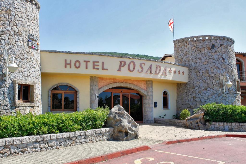 Tējas mājas "Hotel Posada" ieeja ar akmens fasādi un zaļo apstādījumu. Redzama arī karoga mastu ar karogu.