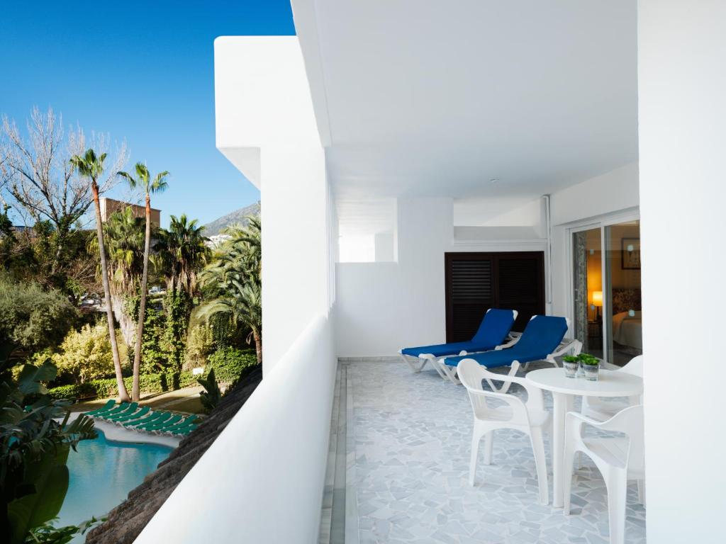 Balconā ir divi zili sauļošanās krēsli un balta galda komplekts, no kura paveras skats uz dārzu ar palmām un baseinu. Gaisa skats ir saulains un skaidrs, radot relaksējošu atmosfēru.