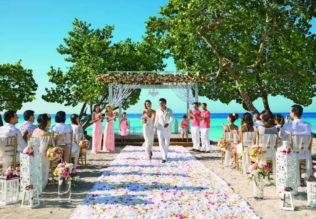 Pludmales kāzu ceremonija ar dekoratīviem ziediem, pāri, kas dodas pa ziediem klātu ceļu, un viesiem, kas svin šos īpašos brīžus zem kokosriekstu kokiem.