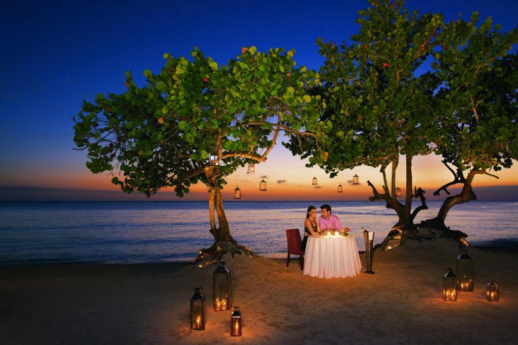 Romantiska vakariņas pie jūras ar sveču gaismu un krāšņu saulrietu. Galda klāšana zem koka, ko apgaismo laternas.