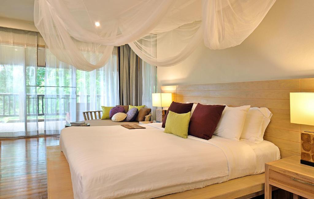 Mūsdienīgs guļamistabas interjers ar plašu gultu, dekoratīvām spilveniem un dabīgu apgaismojumu, kas ieplūst caur lieliem logiem. Gultas virsū ir gaisa tīkls, radot relaksējošu atmosfēru.