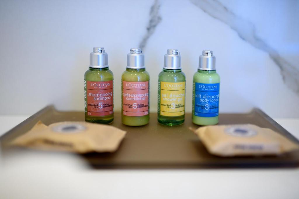 Šajā attēlā redzami L'Occitane zīmola šampūni un ķermeņa kopšanas produkti, kas izvietoti uz pelēka galda. Produkti ietver šampūnu, kondicionieri, dušas želeju un ķermeņa losjonu, kas ir izstrādāti ar dabīgām sastāvdaļām.