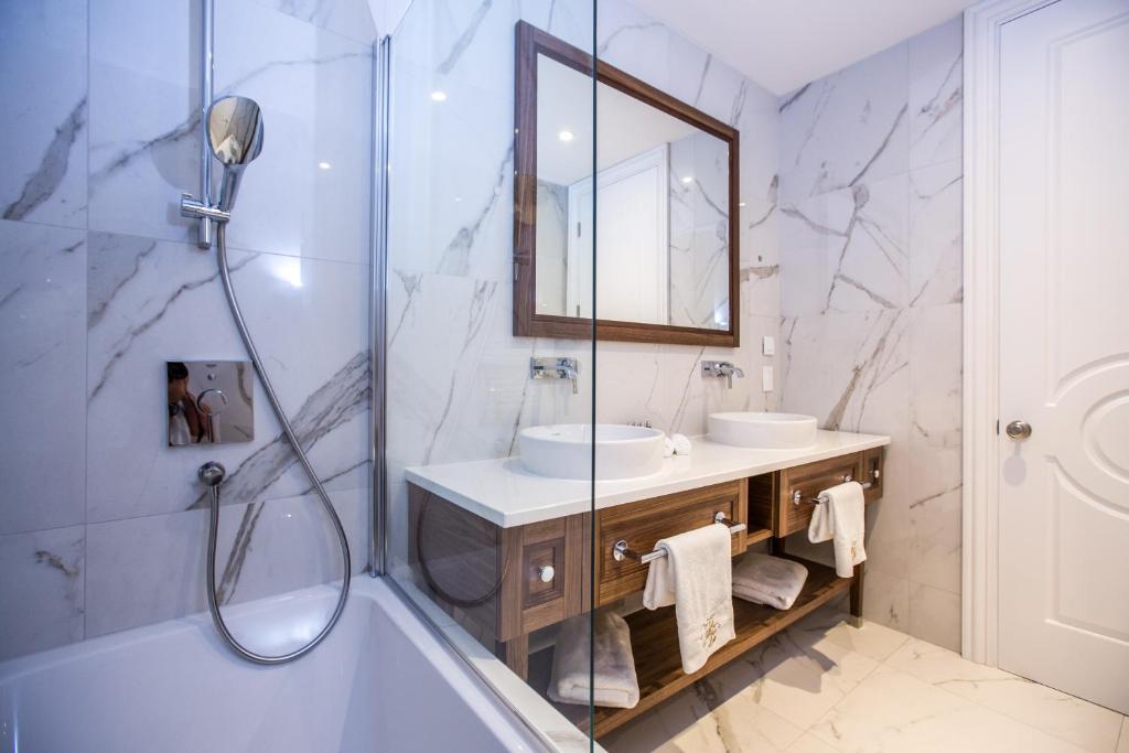 Mūsdienīgs vannas istabas interjers ar marmora apdari, dušas kabīni un divām izlietnēm uz koka skapīša. Vannas istaba ir gaiša un eleganta, radot relaksējošu atmosfēru.