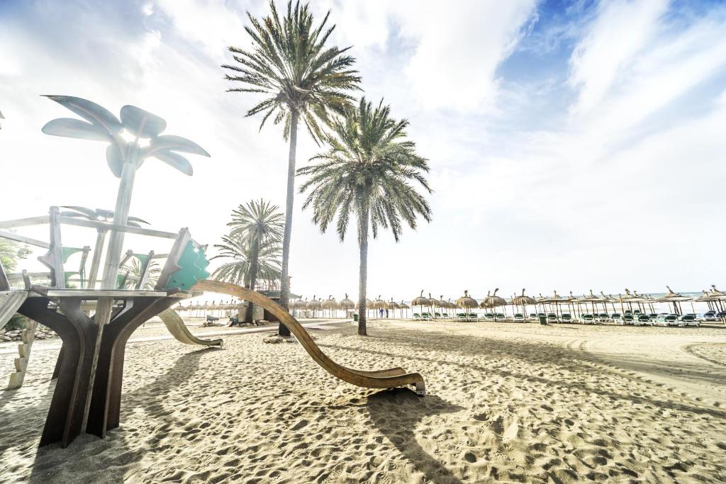 Pludmales rotaļlaukums ar palmām un smiltīm, uz kura redzams slidkalniņš un saulessargu rinda. Ideāla vieta ģimenes atpūtai un bērnu izklaidei.