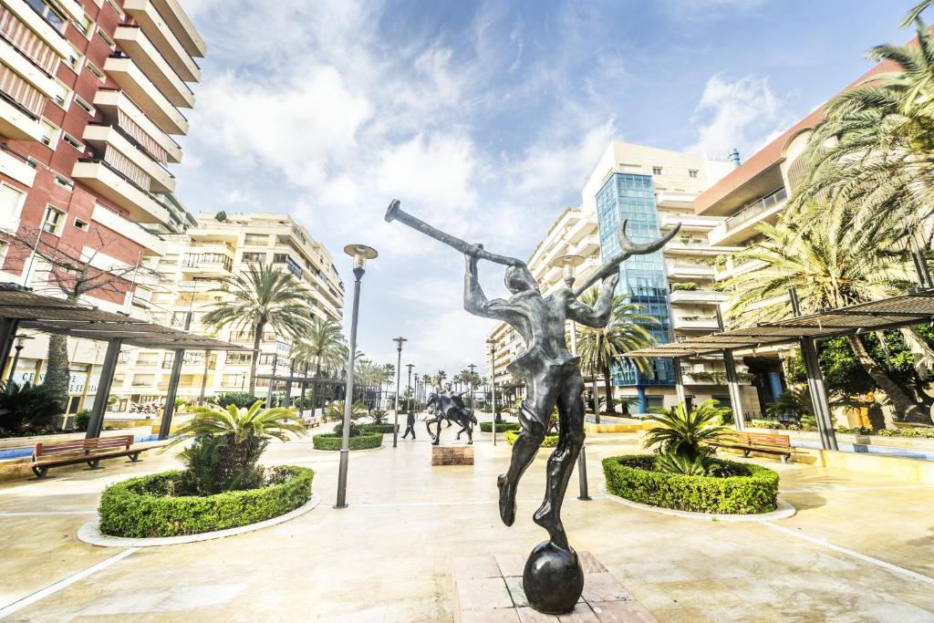 Parks ar skulptūru, kas attēlo cilvēku ar trompeti, apzaļumots un moderns pilsētas ainava ar palmām un ēkām.