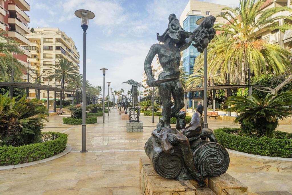 Parks ar bronzas skulptūrām un palmām, kas veido skaistu pilsētas ainavu. Apkārt ir moderni ēkas, un gaisma rada patīkamu atmosfēru.
