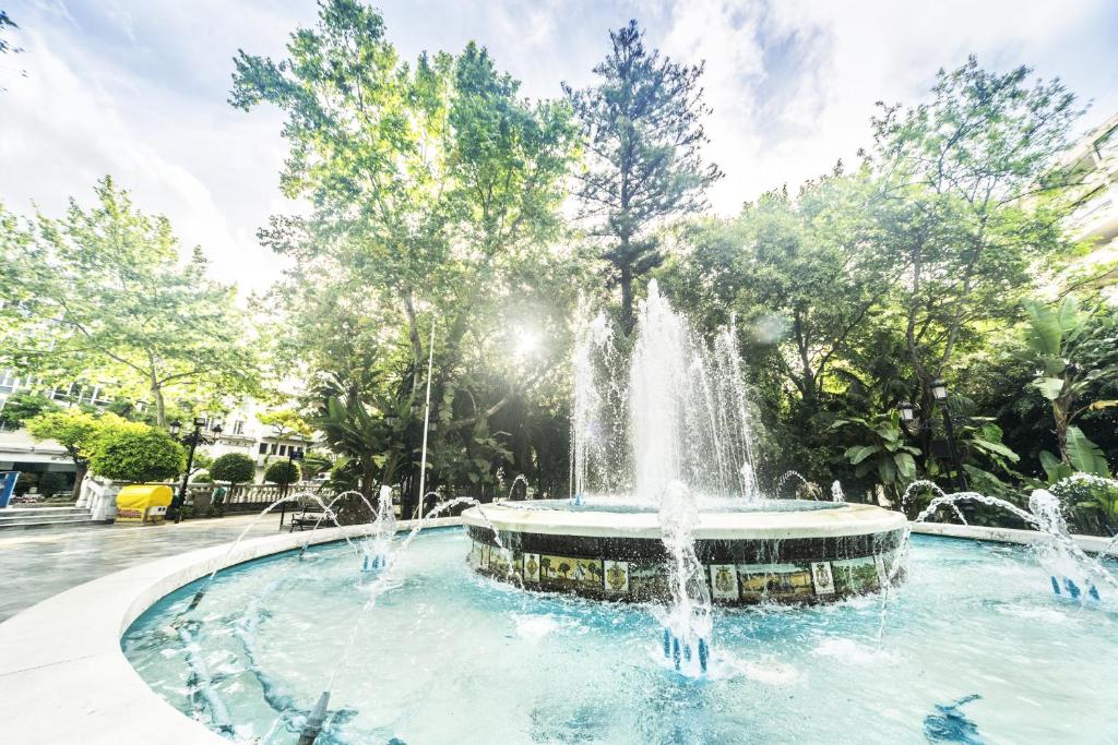 Ūdens strūklaka skaistā parkā starp kokiem, ar saules gaismu, kas caurspīd cauri lapotnei. Mierīga atmosfēra un dabas skaistums.