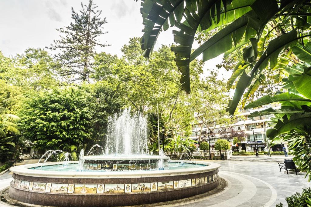 Pūšļa strūklaka zaļajā parkā, ko ieskauj sulīgas augu formas un koku lapas, radot mierīgu un relaksējošu atmosfēru. Fons veido pilsētas ēkas un pastaigu celiņus.