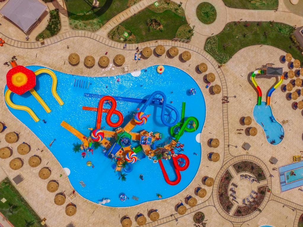 Krāsains ūdens atrakciju parks ar dažādiem slidkalniņiem un peldbaseiniem, kur cilvēki izklaidējās zem saules. Apkārt baseinam redzami sauļošanās krēsli un saules sargiem.