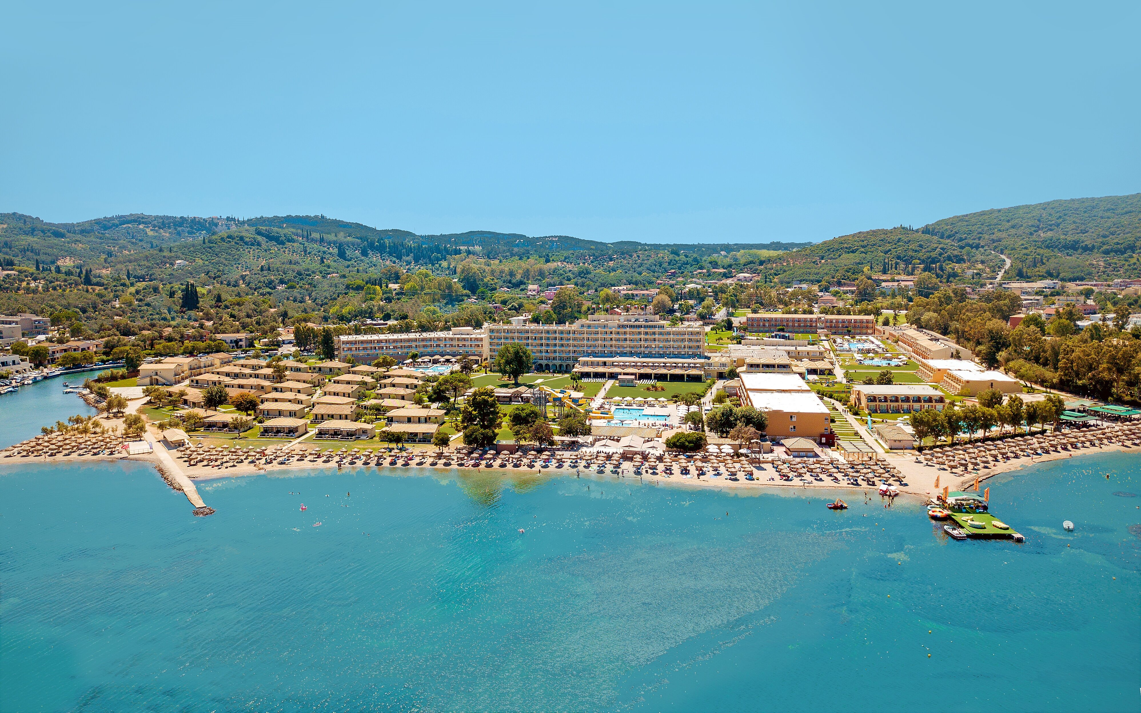 Kūrorta ainava ar pludmali, viesnīcām un zaļām teritorijām, kas atrodas pie ūdens. Skats uz kalniem fona rada mierīgu atmosfēru. panoramic view with beach and pools
