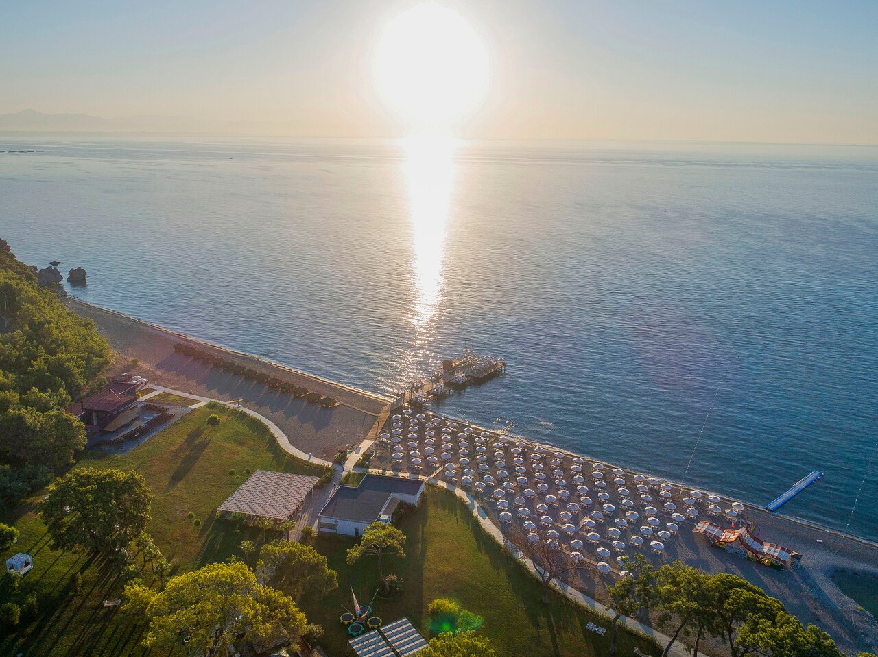 Saulriets virs pludmales ar saulessargiem un piestātni, kas atspoguļojas mierīgajā jūrā. Ainava izceļ skaisto dabu un relaksējošo atmosfēru. panoramic view with beach and pools