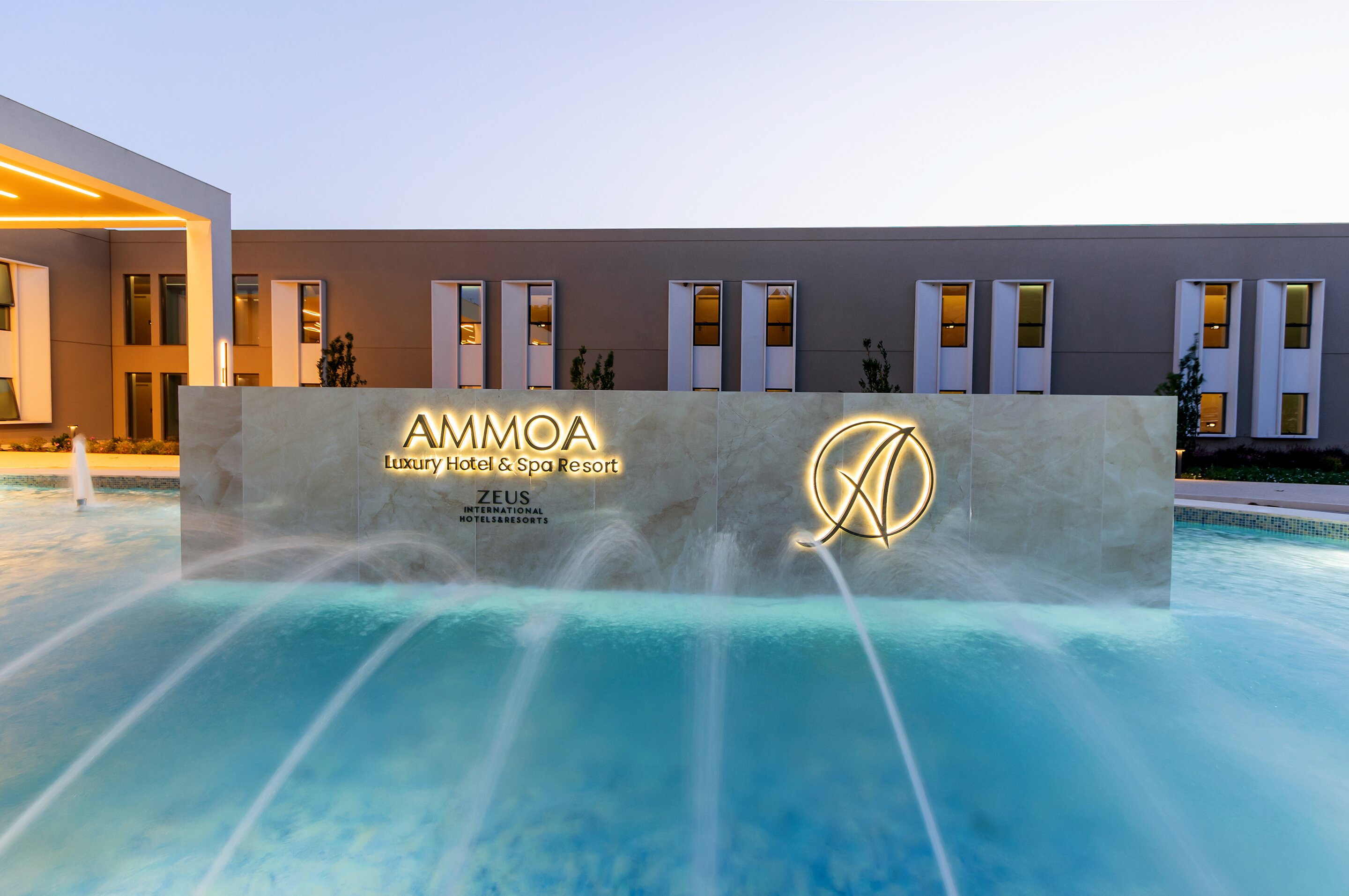 AMMOA luksusa viesnīcas un spa kūrorta izsmalcināta ieeja ar ūdens strūklām, kas rada mierīgu atmosfēru. Izcila zīme ar nosaukumu un logotipu apgaismota vakara laikā. panoramic view with beach and pools