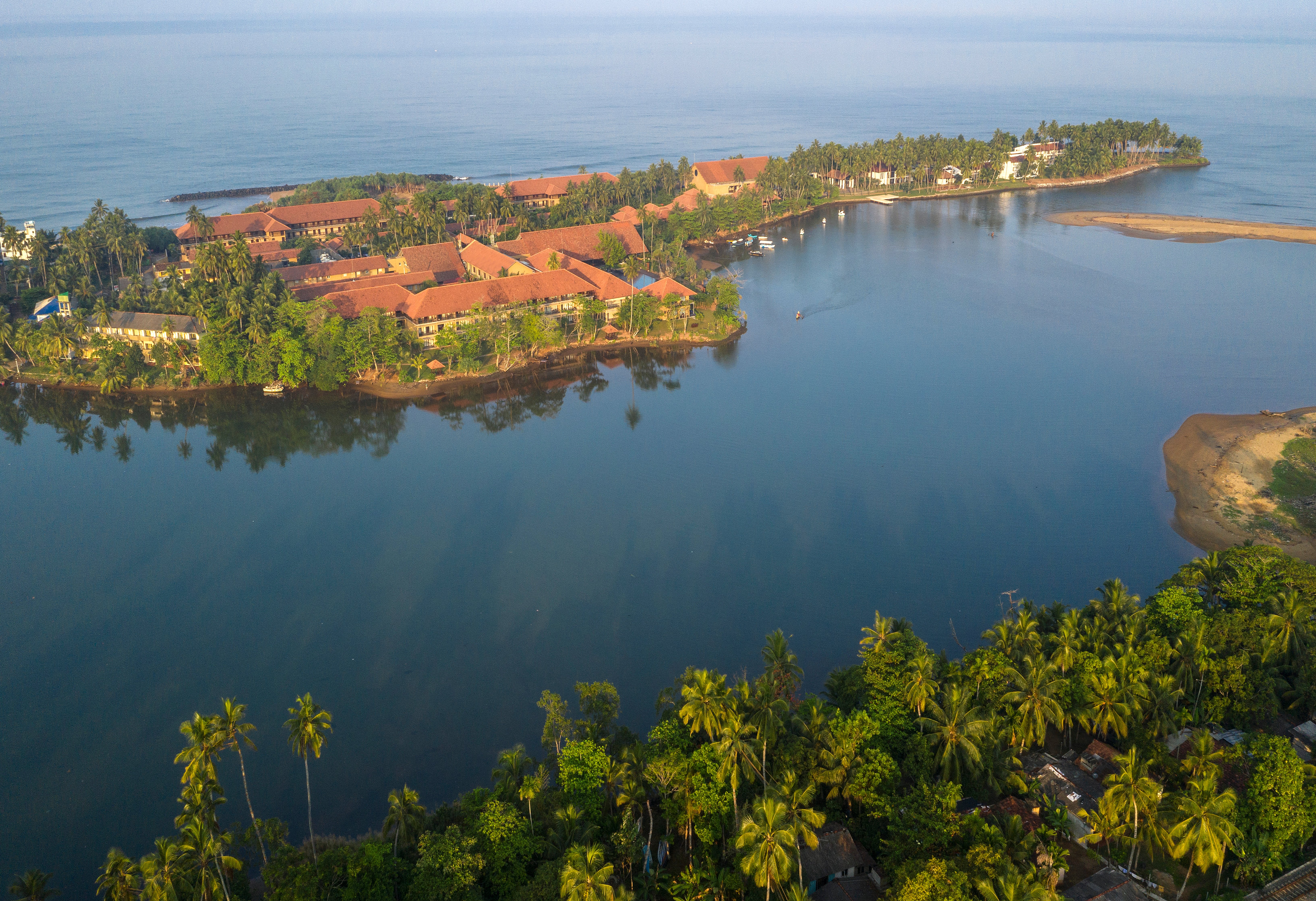 Tropu ainava ar palmām un ūdeni, kur redzams kūrorts pie jūras, kas apvīts ar zaļumiem. Apkaimē ir redzami laivas un smilšaina krasta līnija. panoramic view with beach and pools