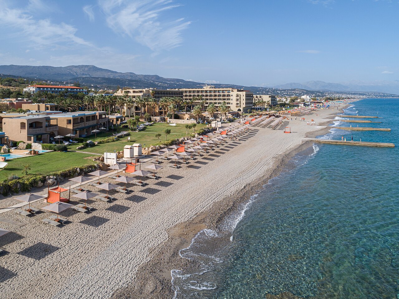 Pludmales ainava ar viesnīcām un saulessargiem, kas izvietoti pie jūras, radot relaksējošu atmosfēru. Zilais ūdens un kalnu fons papildina ainavu. panoramic view with beach and pools