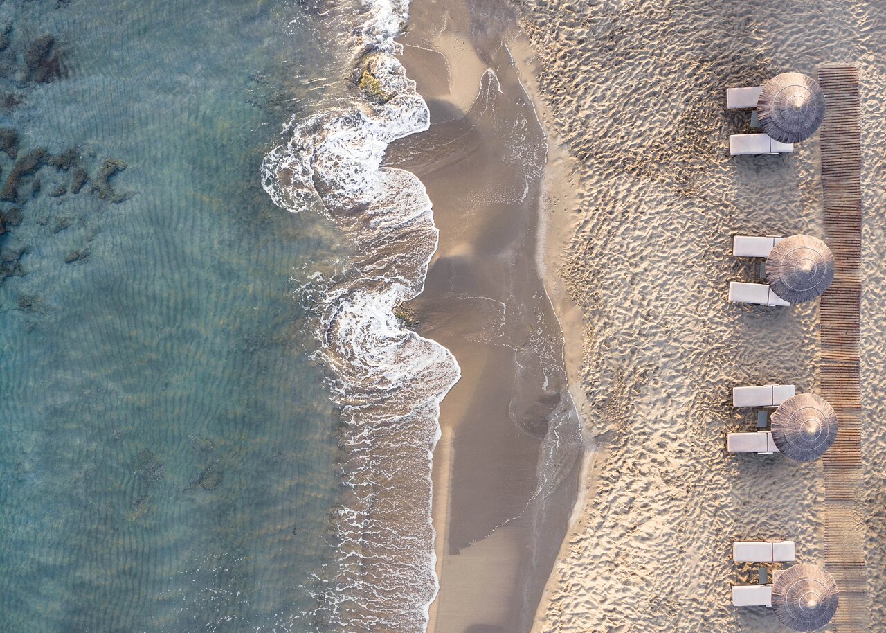 Pludmales ainava no putna lidojuma, kur redzama smilšu krasta līnija un viļņi, kas skar krastu, blakus izvietoti sauļošanās krēsli un salmu saulessargi. panoramic view with beach and pools