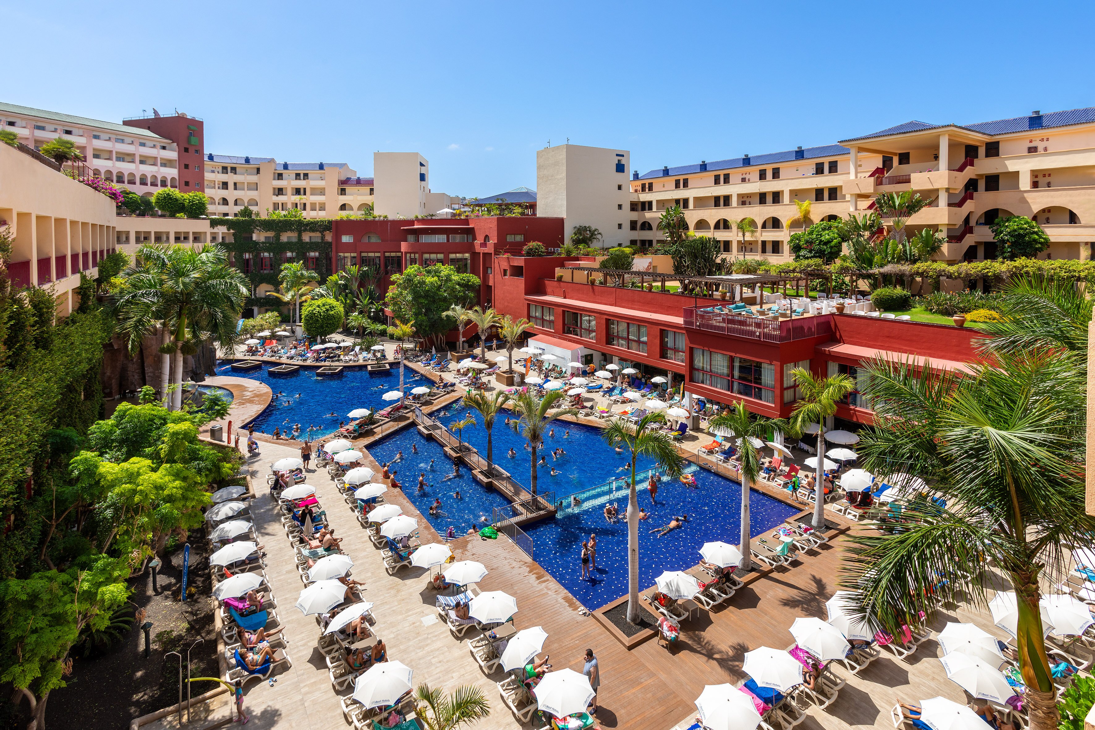 Peldbaseins viesnīcas pagalmā ar saulessargiem un atpūtniekiem, apkārt palmām un apzaļumojumam. Skats uz ēkām un zilo debesu fona. panoramic view with beach and pools