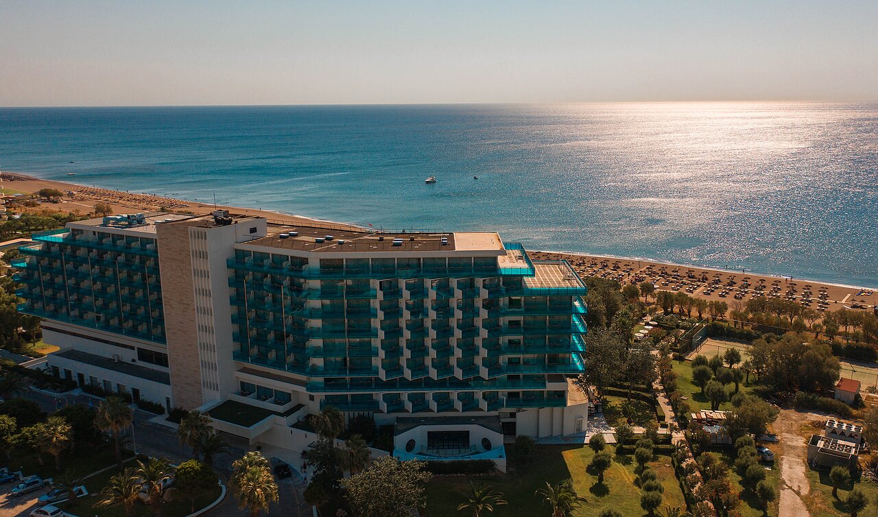 Mūsdienīgs viesnīcas komplekss blakus pludmalei, ar skatu uz jūru un saulrietu. Apkārtne ir zaļa ar palmām un sauļošanās krēsliem. panoramic view with beach and pools