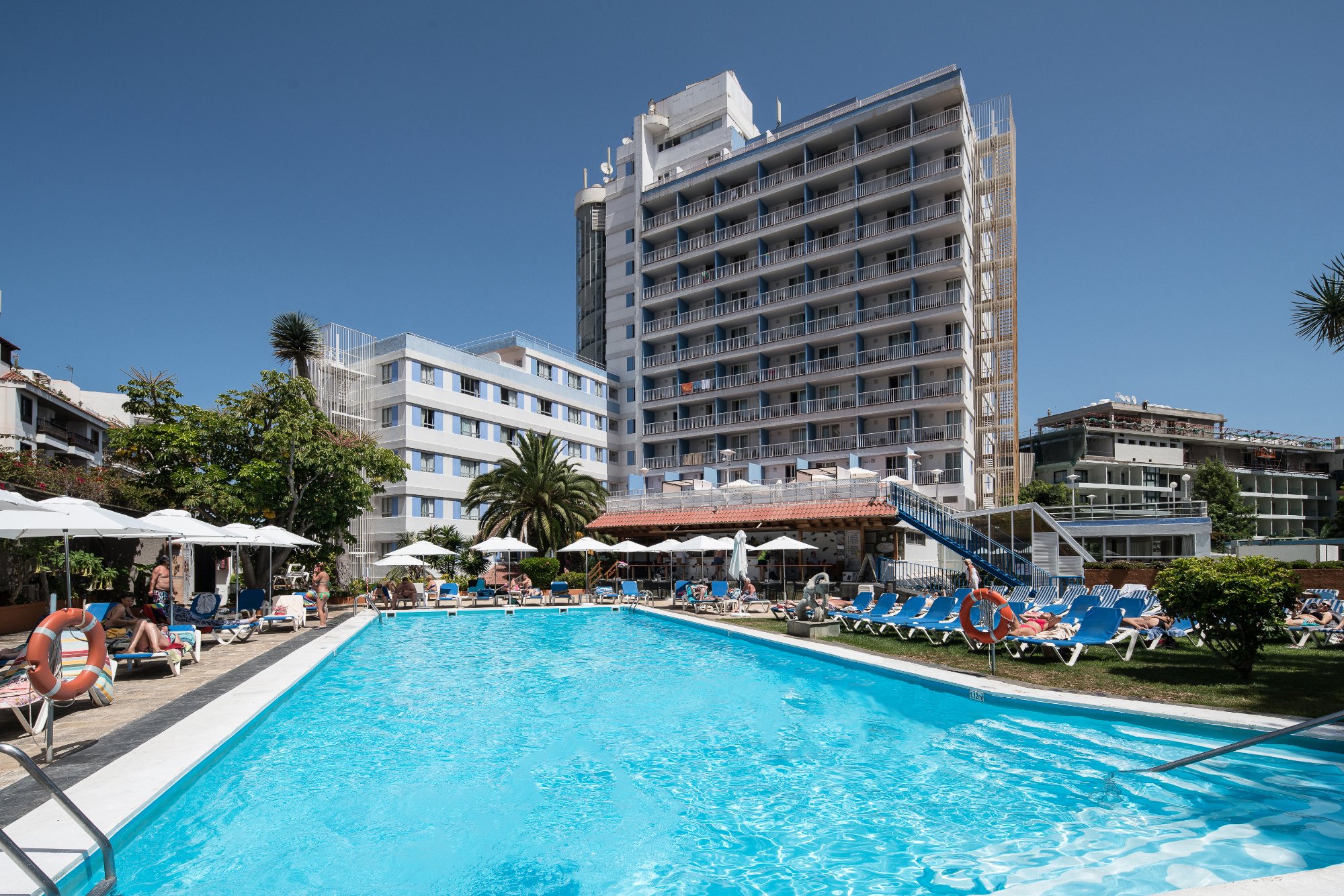 Baseins ar skaidru ūdeni, apkārt esošās viesnīcas un atpūtnieki, kas relaksējas saulē. Viesnīcas fasāde ir moderna, ar balkoniem un baseina zonu. panoramic view with beach and pools
