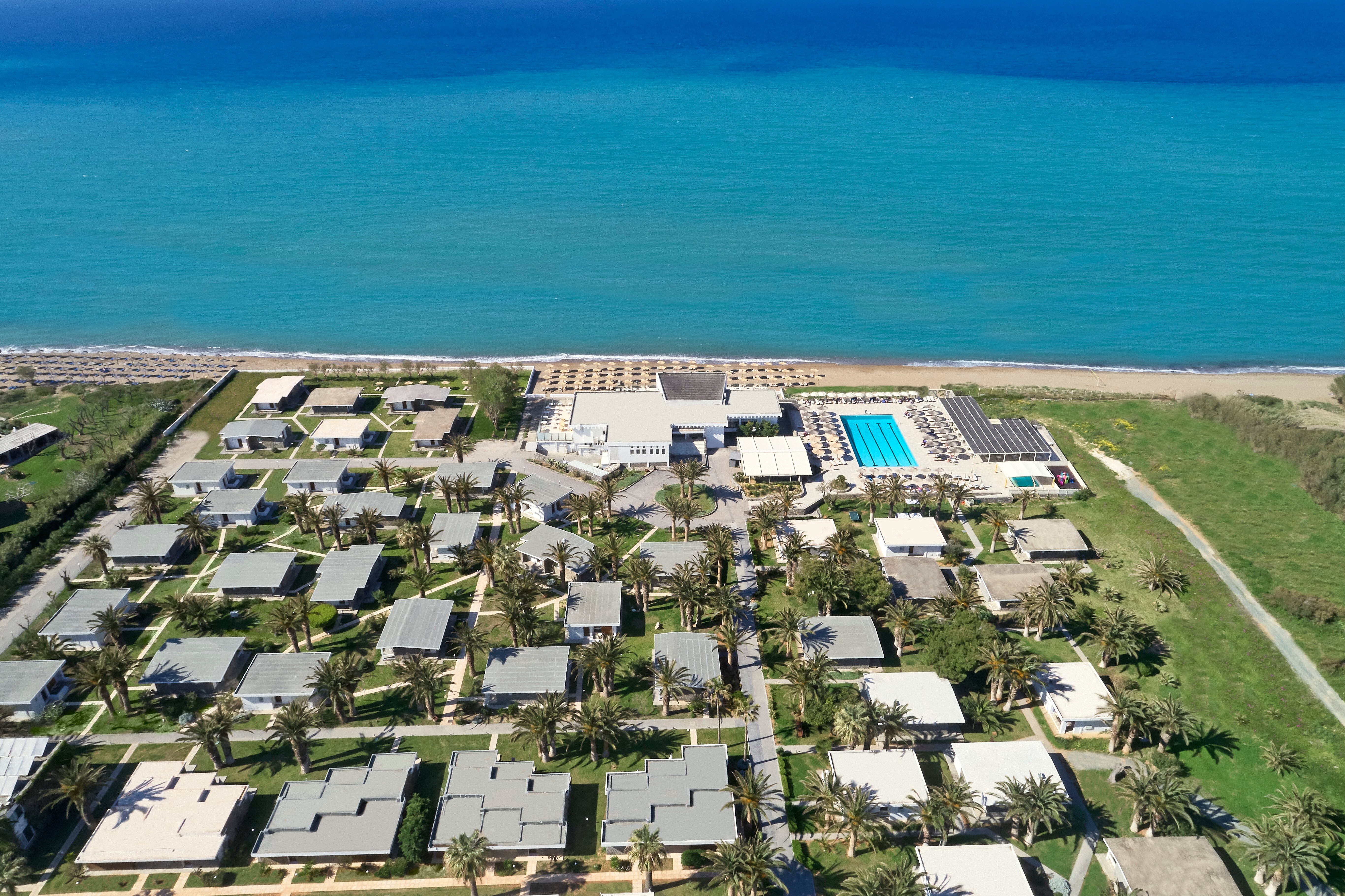 Kūrorts pie jūras ar viesnīcu kompleksu un baseinu, ieskauts zaļām palmu alejām. Jūras krasts redzams fonā, sniedzot relaksējošu atmosfēru. panoramic view with beach and pools