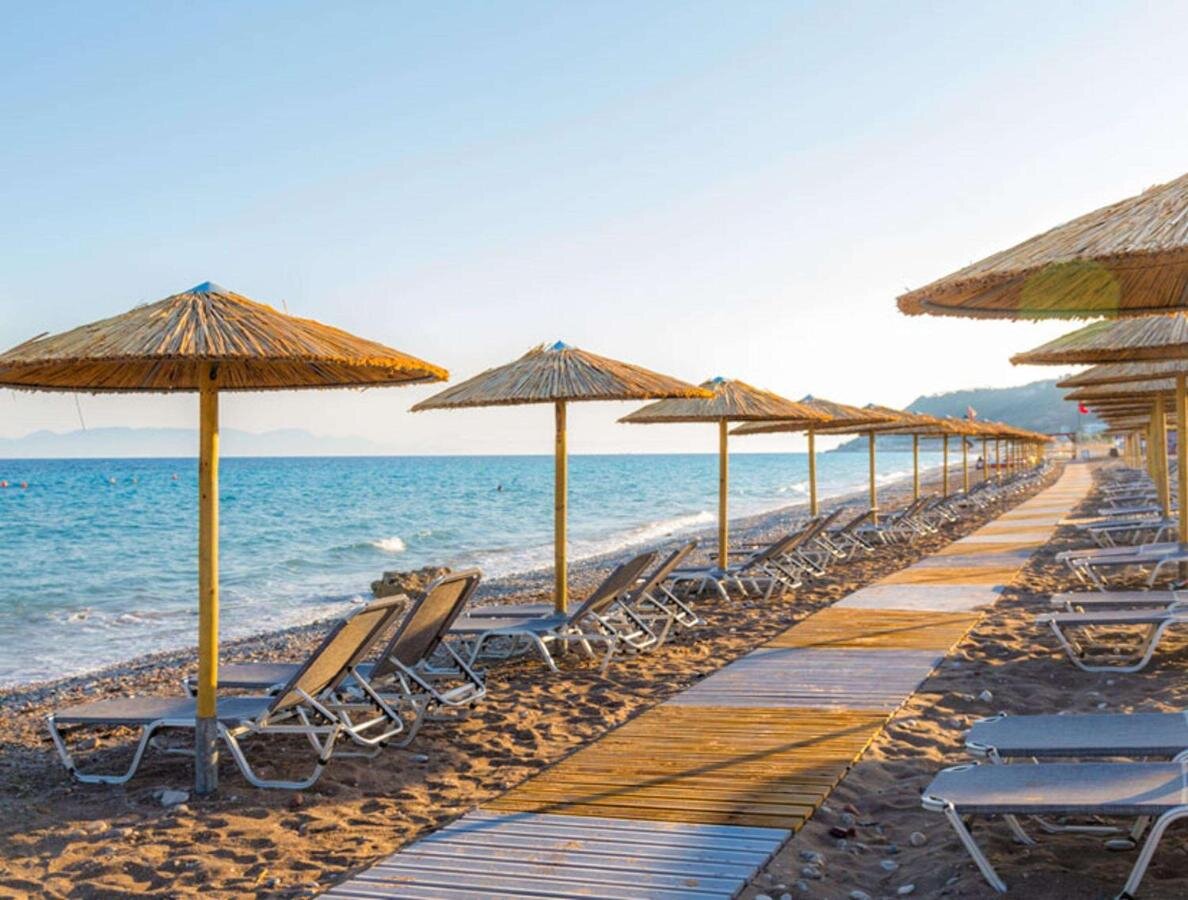 Pludmales ainava ar sauļošanās krēsliem un salmiem jumtiem, kas izvietoti gar smilšu krastu. Zilās jūras viļņi un skaidra debess veido mierīgu atmosfēru. panoramic view with beach and pools