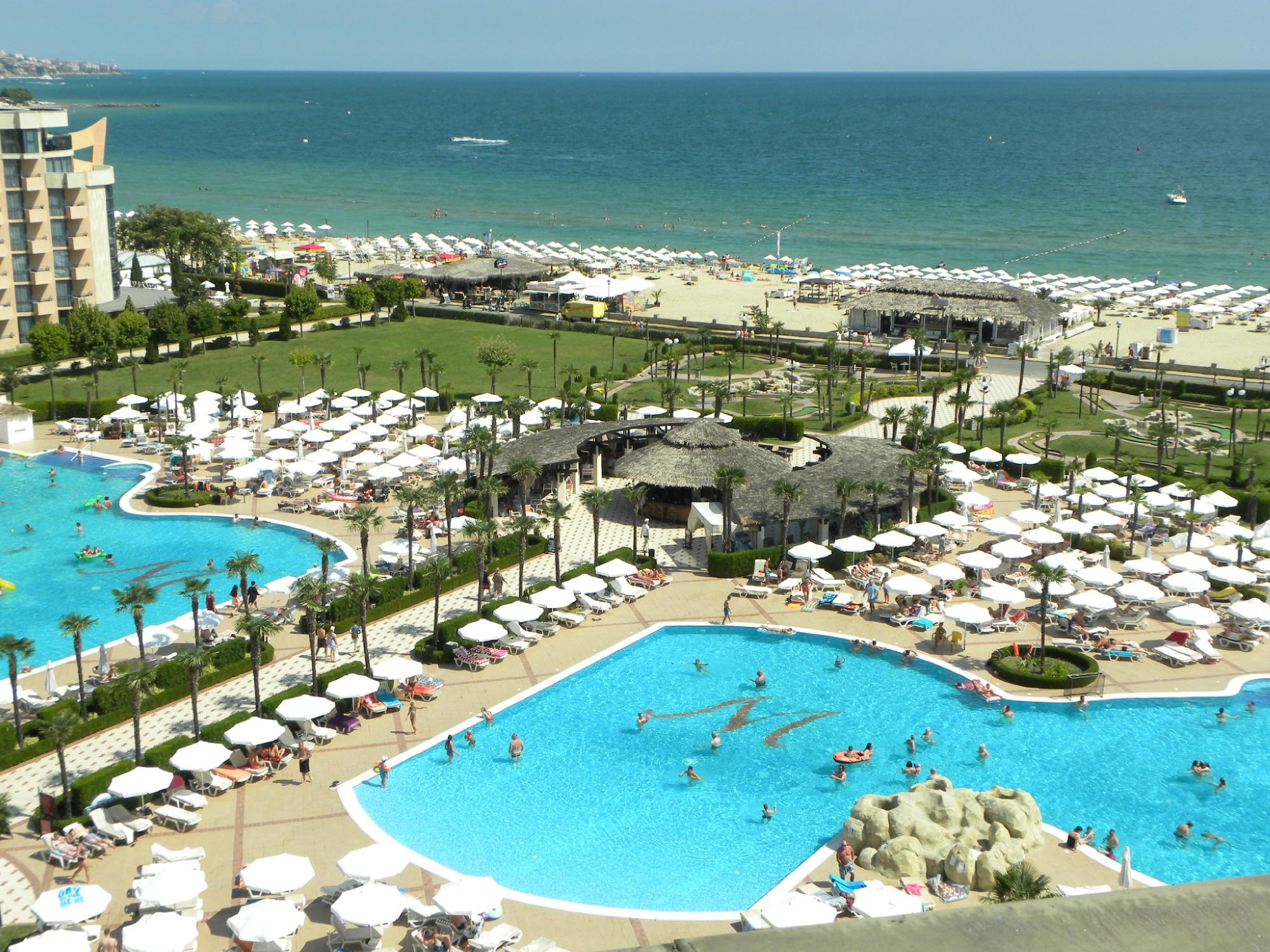 Vasaras kūrorts ar peldbaseiniem un pludmali, kur cilvēki atpūšas zem saules. Skaista jūras ainava un zaļie zālieni pie viesnīcas. panoramic view with beach and pools