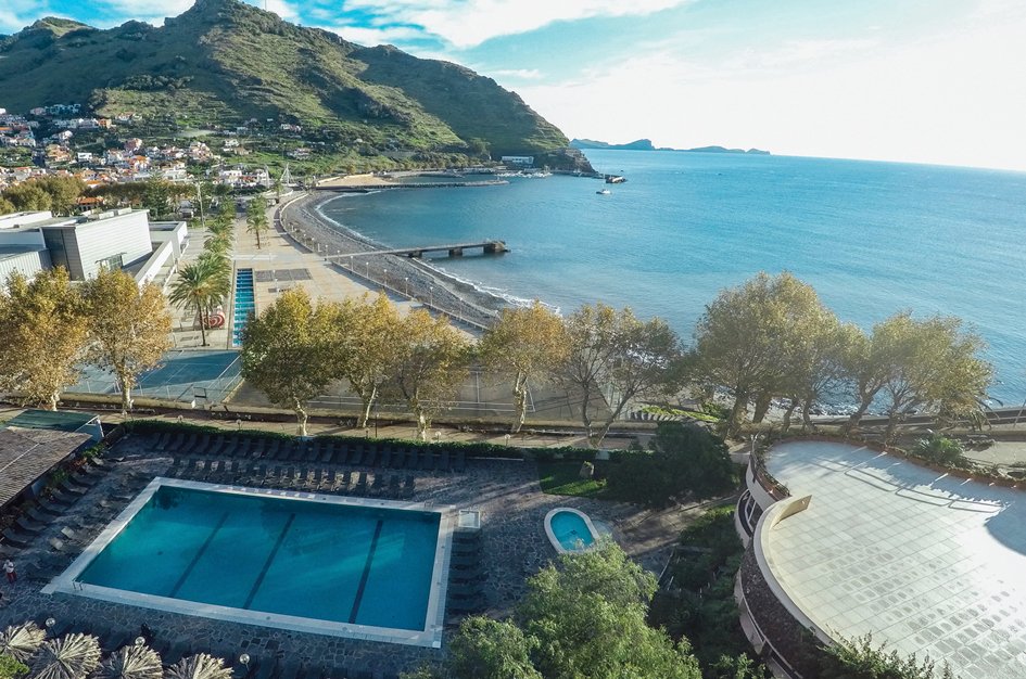 Skats uz pludmali ar kalniem fonā, kur redzams baseins un promenāde, kas ved uz jūru. Apkārtnē ir koki un ēkas, kas veido gleznainu ainavu. panoramic view with beach and pools