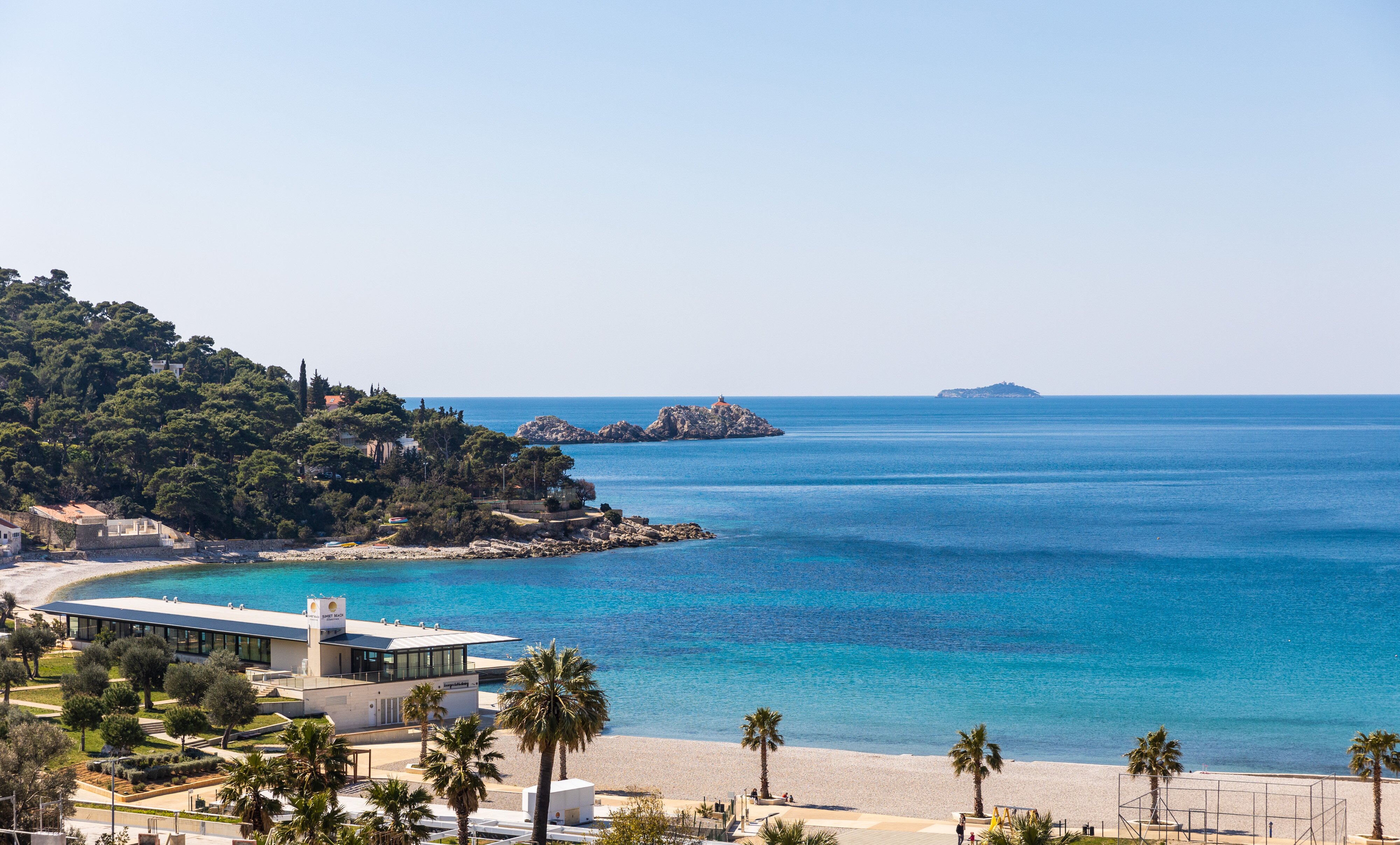 Jūras ainava ar skaidru zilu ūdeni, pludmali un zaļiem kokiem kalnos, kas sniedz mierīgu atmosfēru. Redzami akmeņi jūrā un moderna ēka pludmalē. panoramic view with beach and pools