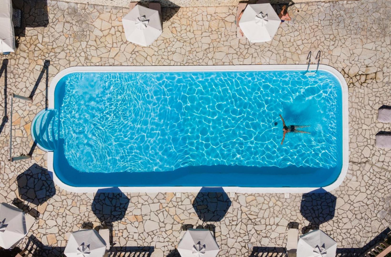 Aurora Beach Hotel āra baseins no putna lidojuma Korfu salā — dzidri zils ūdens, akmens flīžu klājums un balti saulessargi.