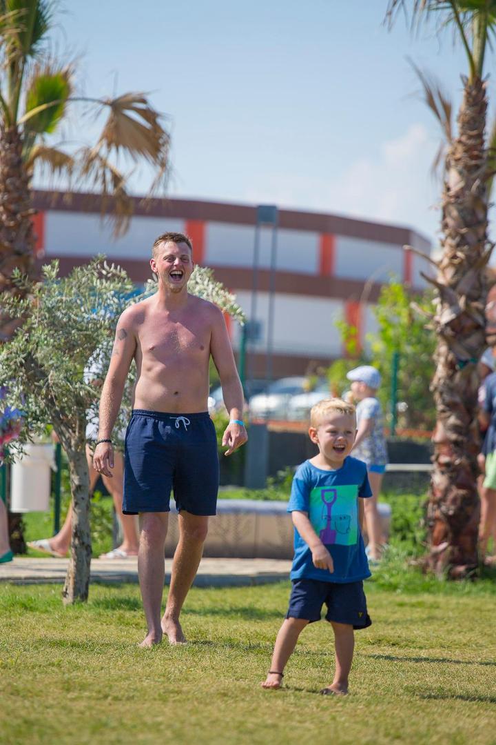Tēvs un dēls smaida un skrien pa zālienu Numa Bay Exclusive Hotel teritorijā Alanjā, fonā palmas
