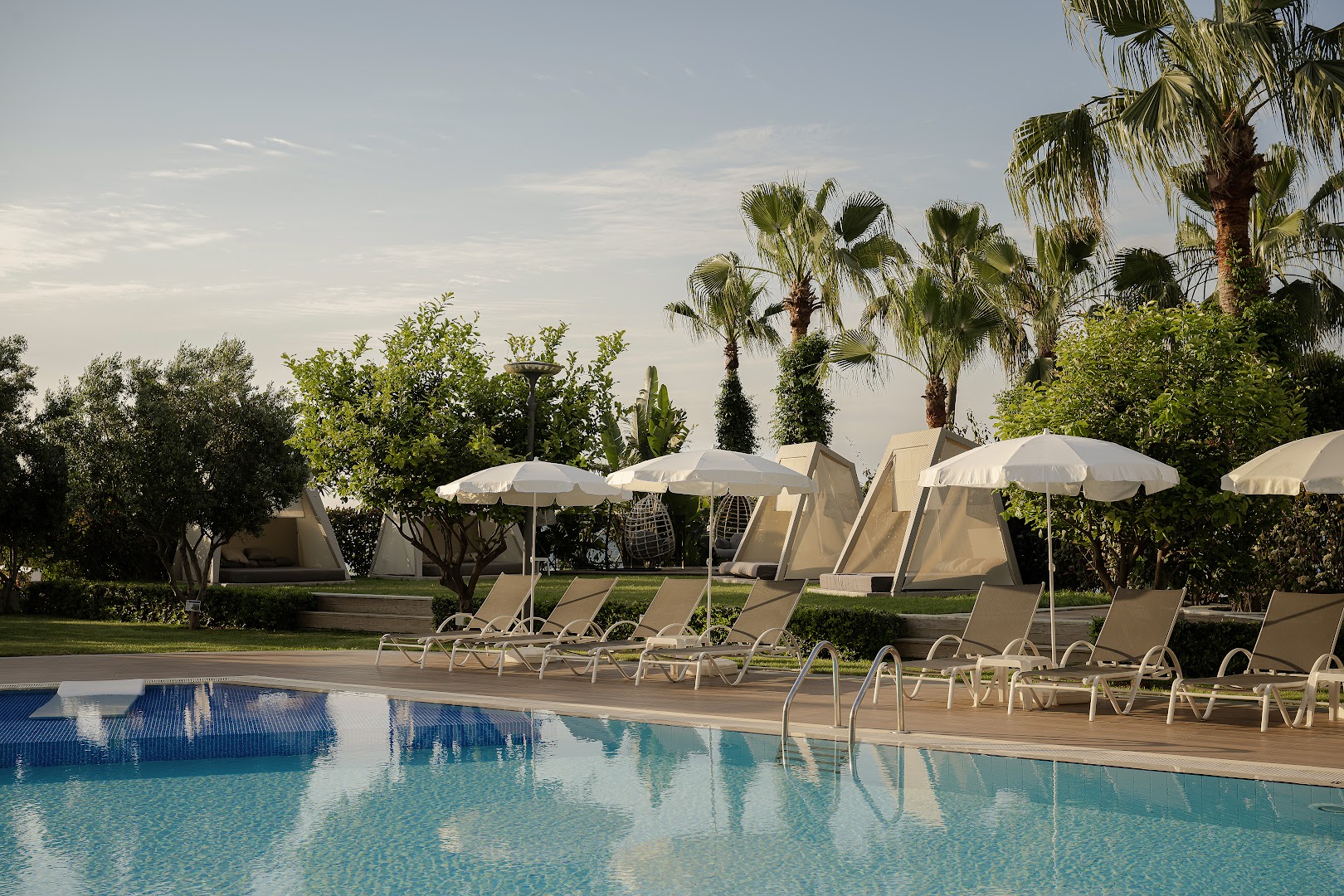 Selene Beach and Spa Hotel āra baseins ar sauļošanās krēsliem, baltiem saulessargiem un palmām fonā — plaša atpūtas teritorija saulainā dienā