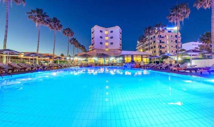 Pavlo Napa Beach Hotel ēka un izgaismots āra baseins nakts laikā Ajanapā — palmas un sauļošanās krēsli apkārt