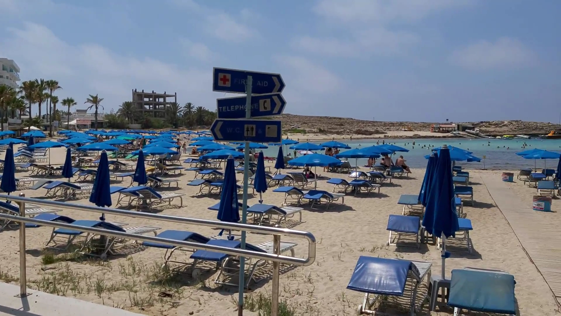 Pavlo Napa Beach Hotel smilšu pludmale Ajanapā, Kiprā — skats uz ziliem saulessargiem, sauļošanās krēsliem un norādēm pie jūras krasta
