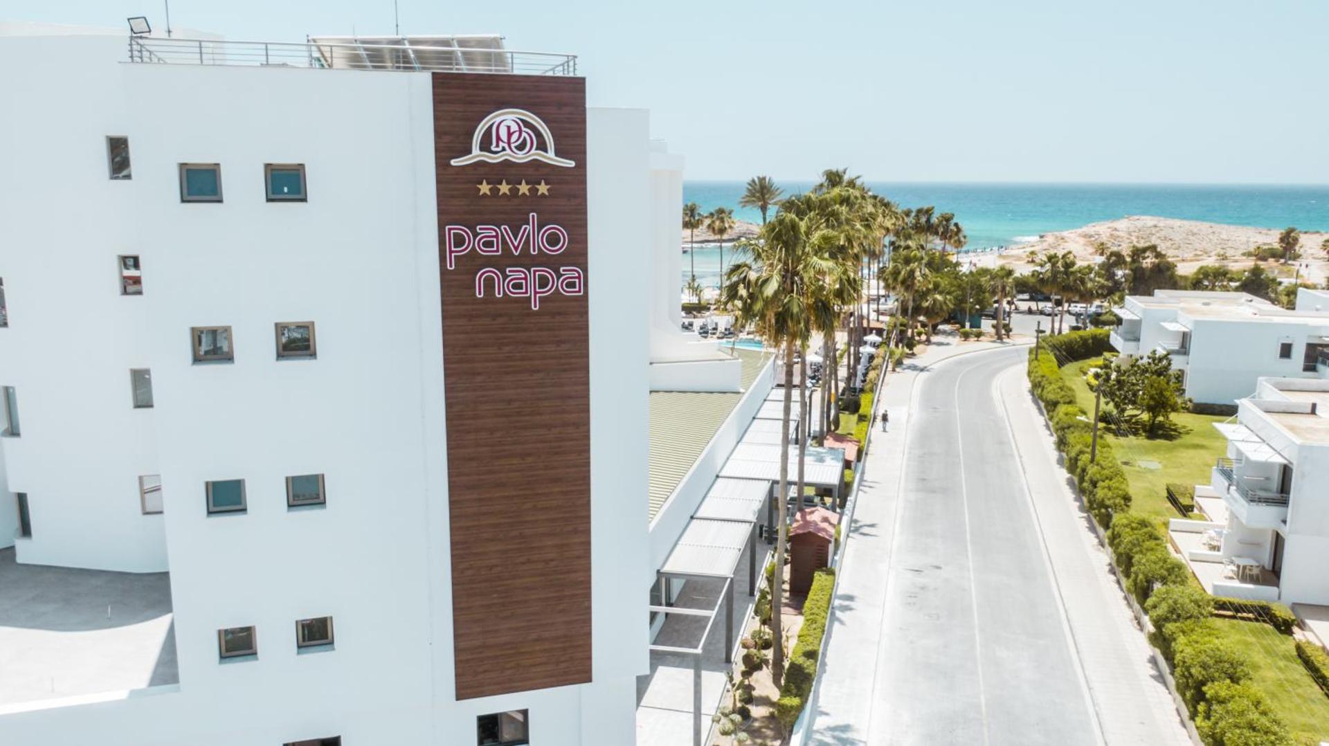 Pavlo Napa Beach Hotel 4* viesnīcas fasāde ar logo Ajanapā — skats uz piebraucamo ceļu, palmām un Vidusjūras piekrasti tālumā