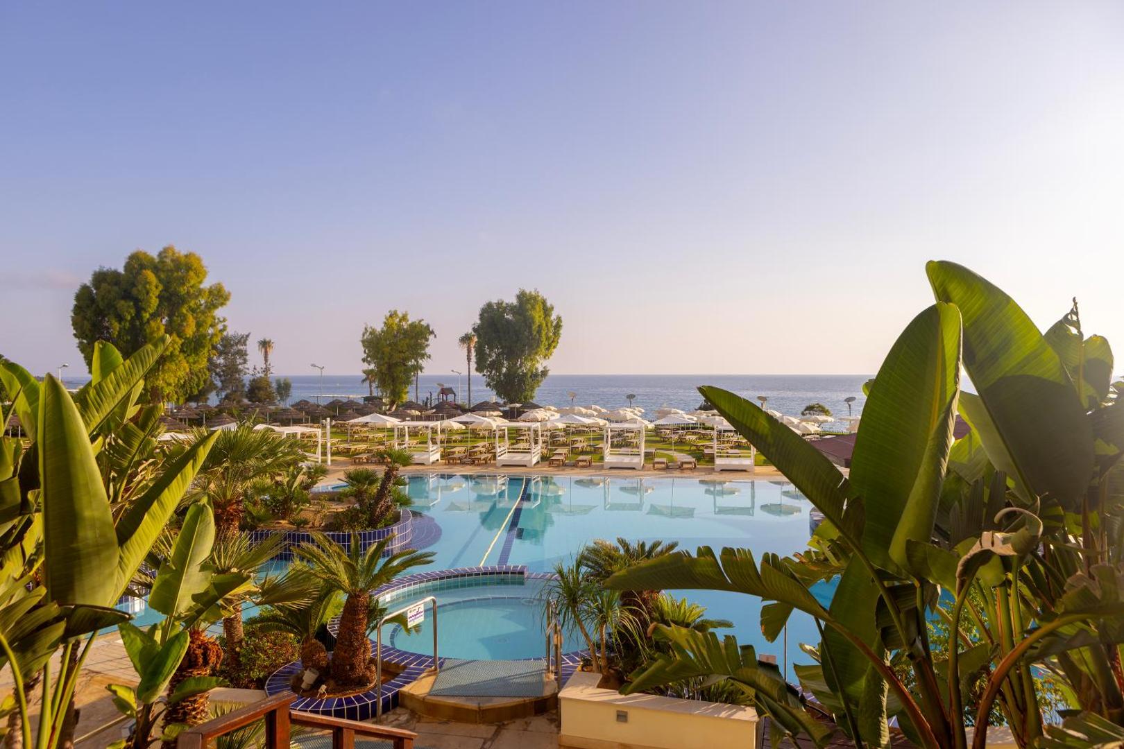 Capo Bay Beach Hotel plašais āra baseins ar palmām un eksotiskiem augiem, fonā Vidusjūra un saulessargu zona Protarasā