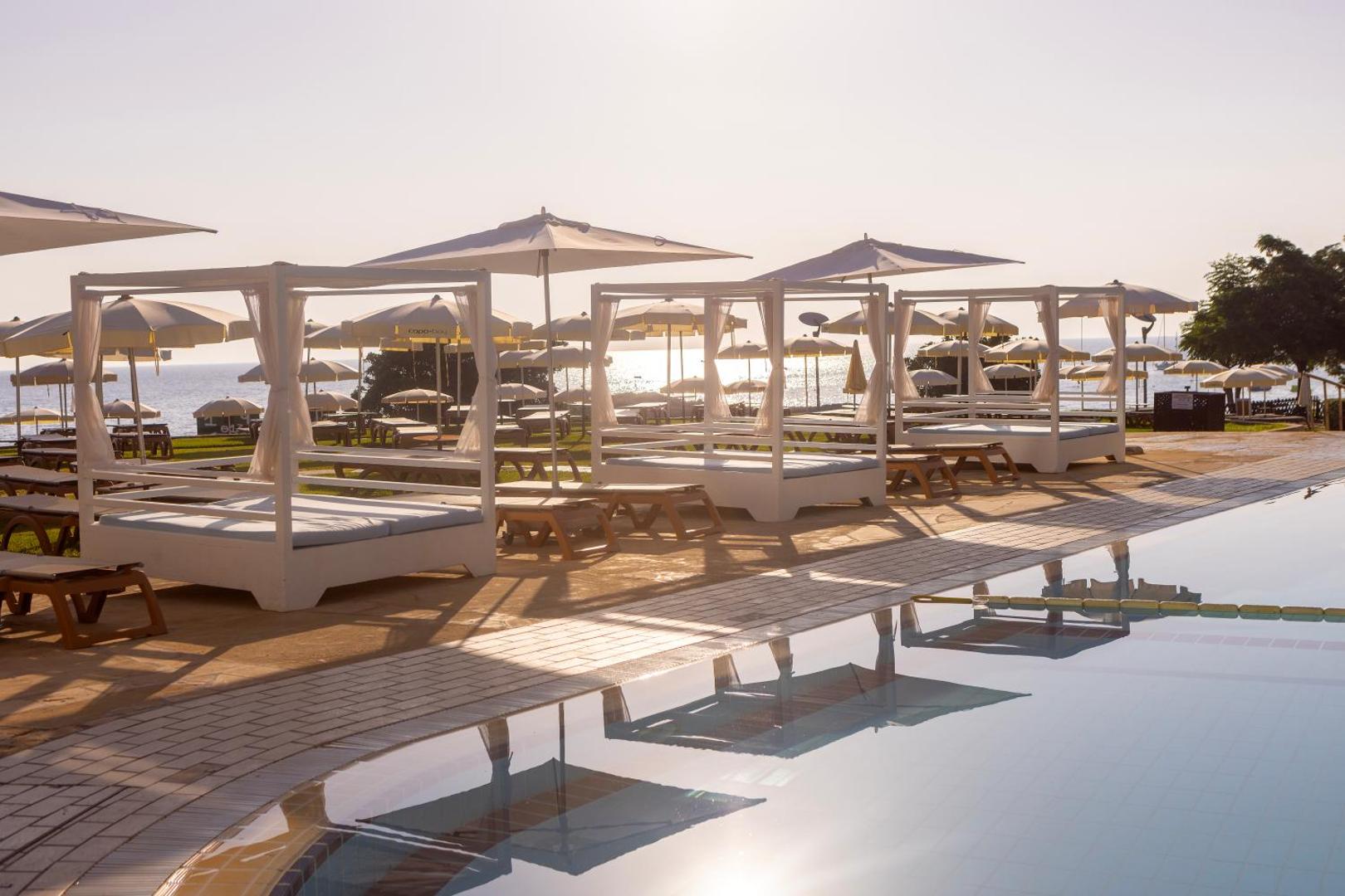 Capo Bay Beach Hotel atpūtas zona pie baseina ar baltām gultām un saulessargiem saulrieta gaismā pie jūras krasta