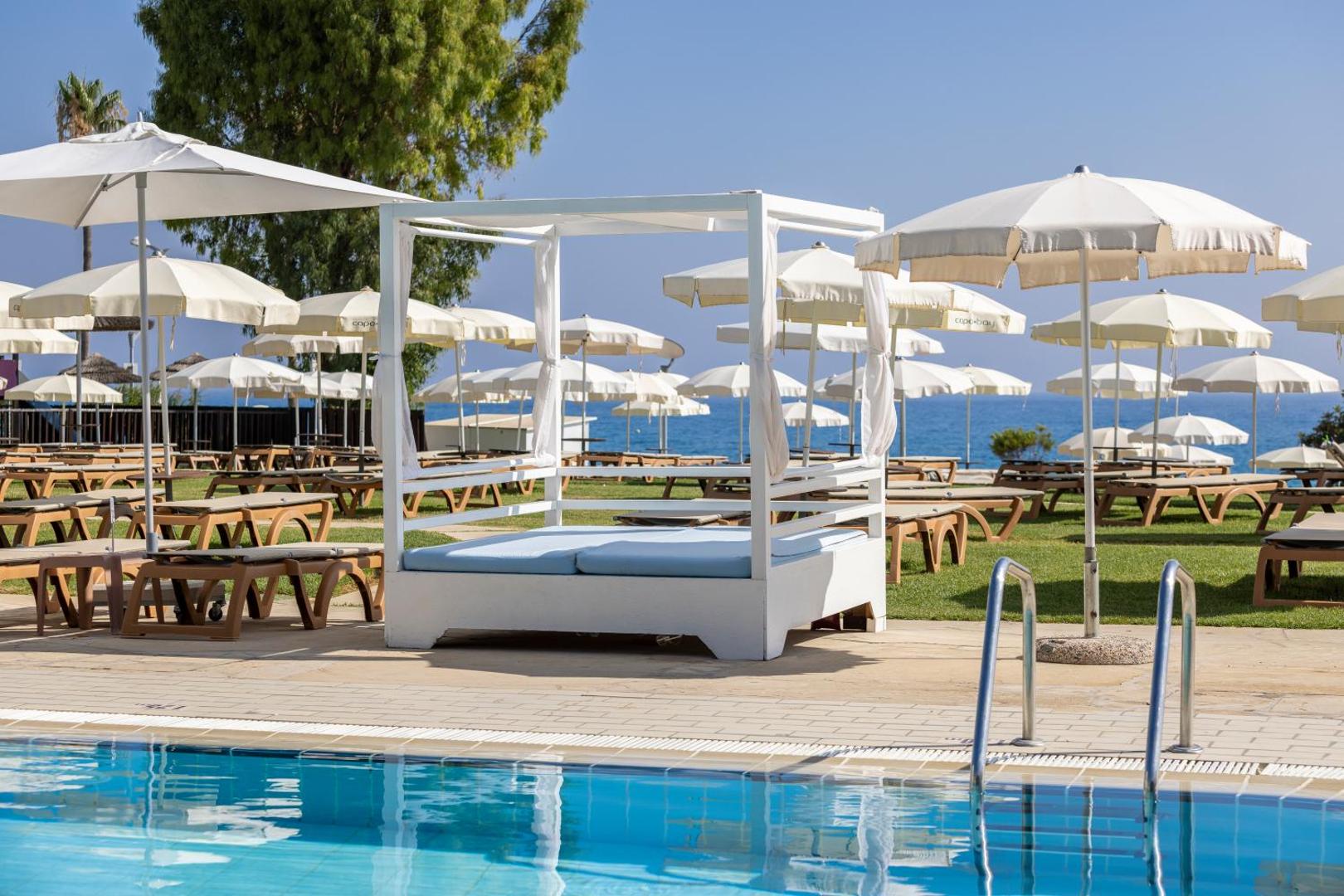 Capo Bay Beach Hotel atpūtas zona ar baltu baldahīnu un saulessargiem pie baseina Protarasā, Kiprā — skats uz jūru un zālienu