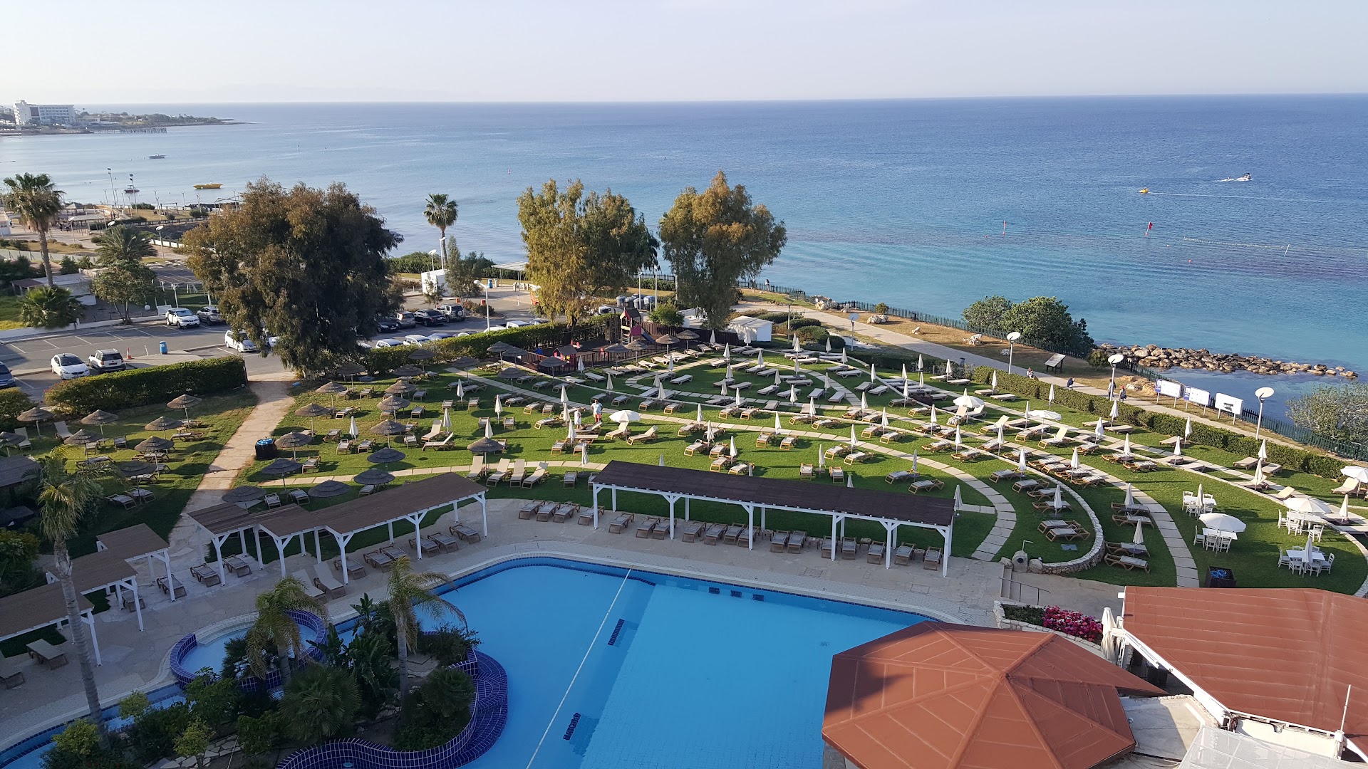 Skats no augšas uz Capo Bay Beach Hotel teritoriju — baseins, zālājs ar sauļošanās krēsliem un Vidusjūras piekraste fonā