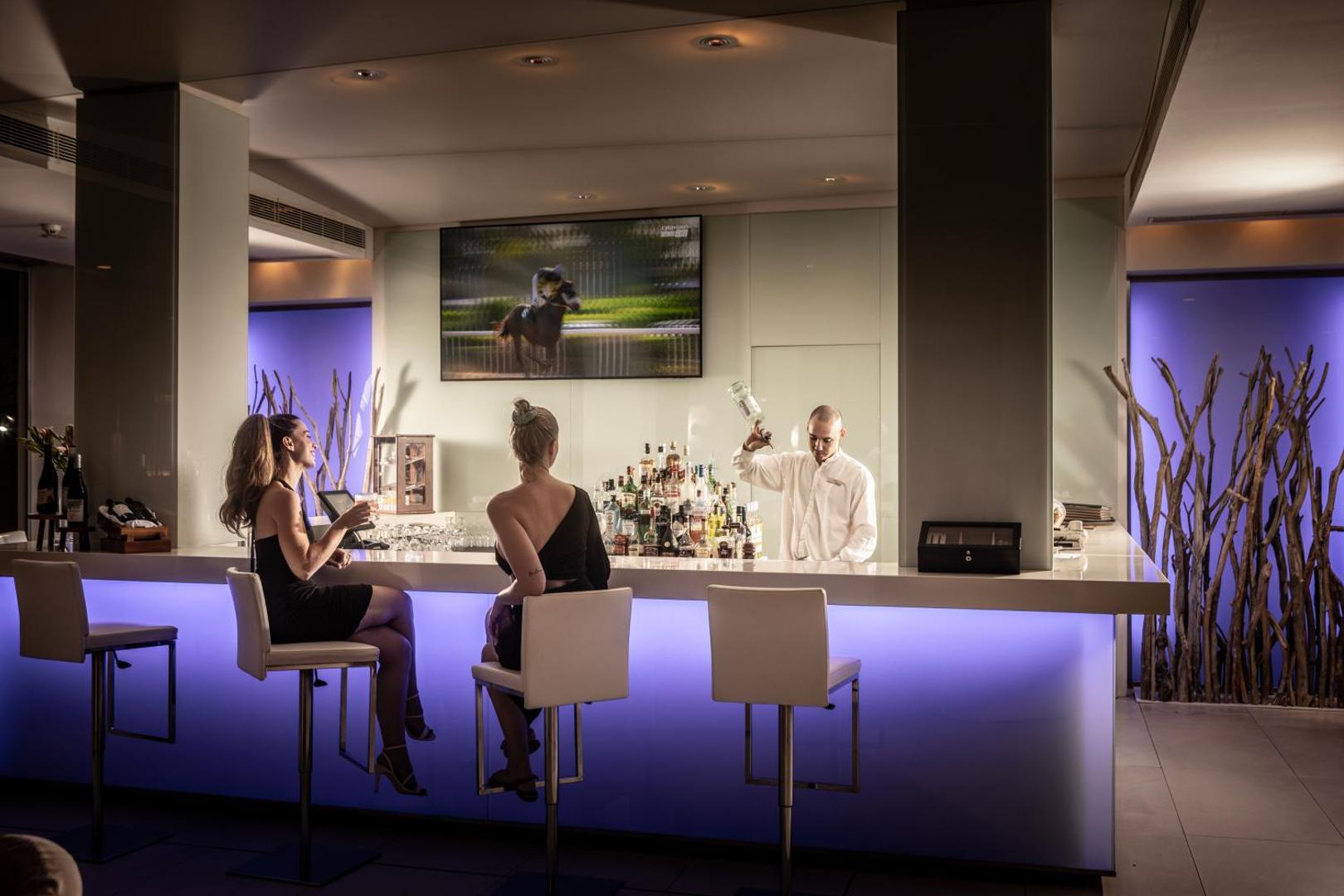 Capo Bay Beach Hotel modernais lobby bārs ar zilu apgaismojumu un bārmeni, kurš gatavo kokteiļus viesiem vakarā Protarasā, Kiprā