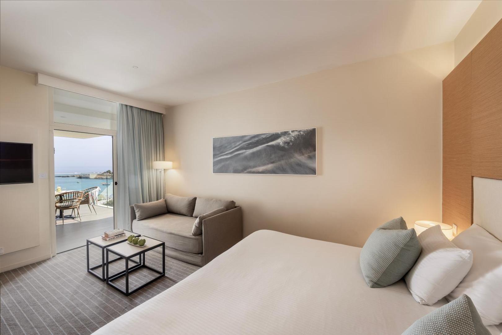 Viesnīcas Capo Bay Beach Hotel numura interjers ar dīvānu un skatu uz balkonu, no kura paveras Vidusjūra