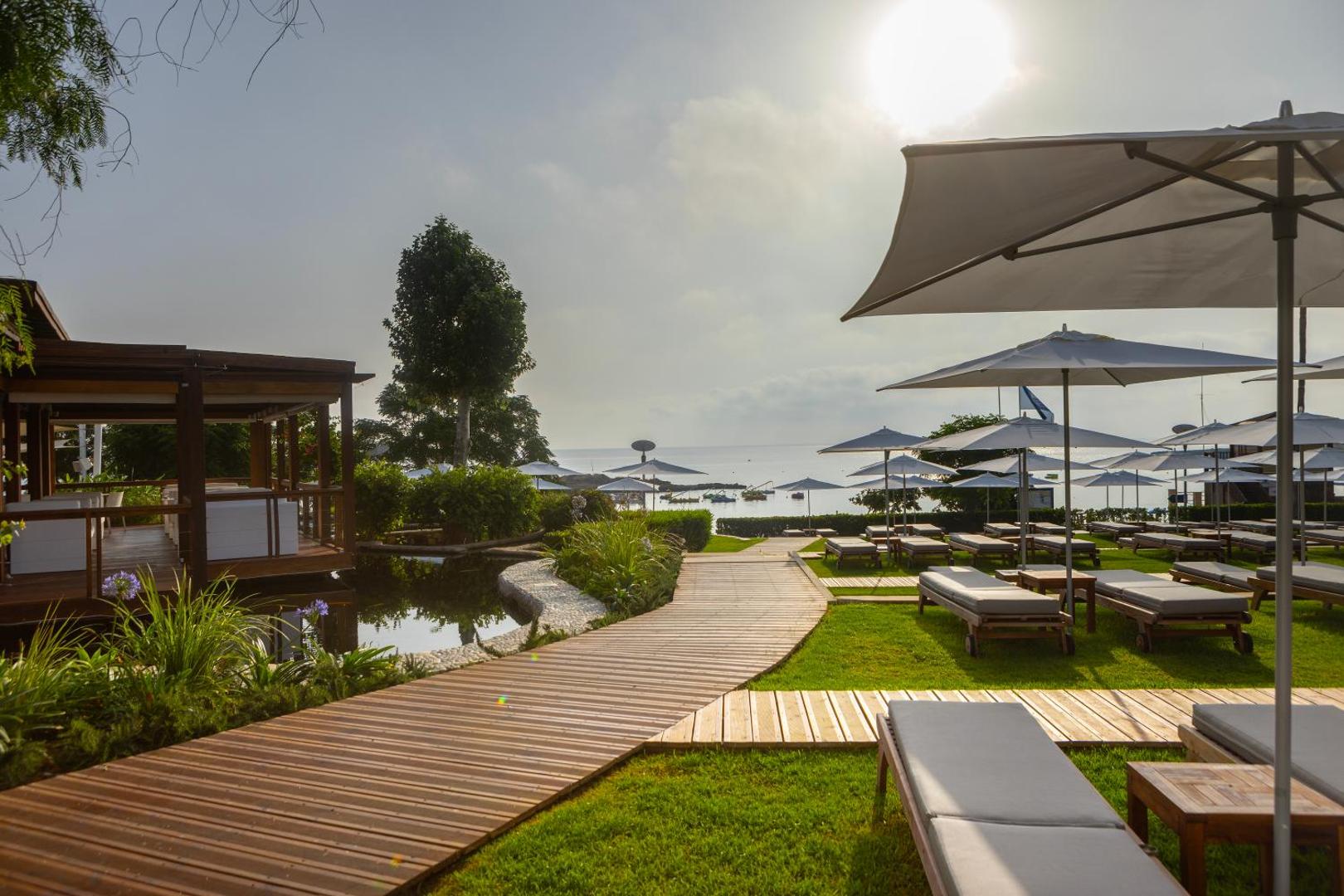 Capo Bay Beach Hotel dārza zona ar koka laipām, zālāju un sauļošanās krēsliem zem saulessargiem pie Protarasas piekrastes