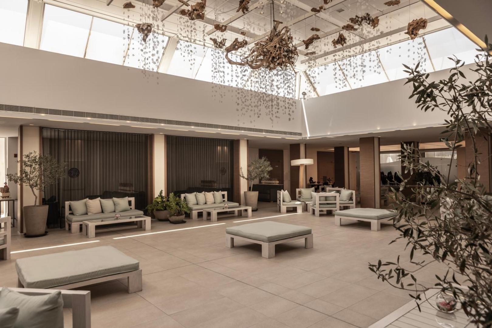 Capo Bay Beach Hotel plašais vestibils ar stikla jumtu, dizaina gaismekļiem un gaišām mēbelēm atpūtai