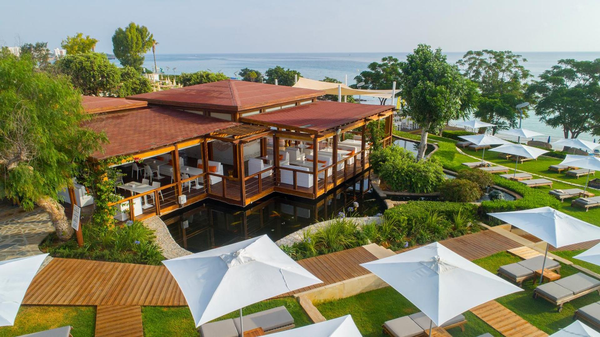 Capo Bay Beach Hotel restorāna terase virs dekoratīva dīķa ar skatu uz jūru un sauļošanās zonu zālienā Protarasā