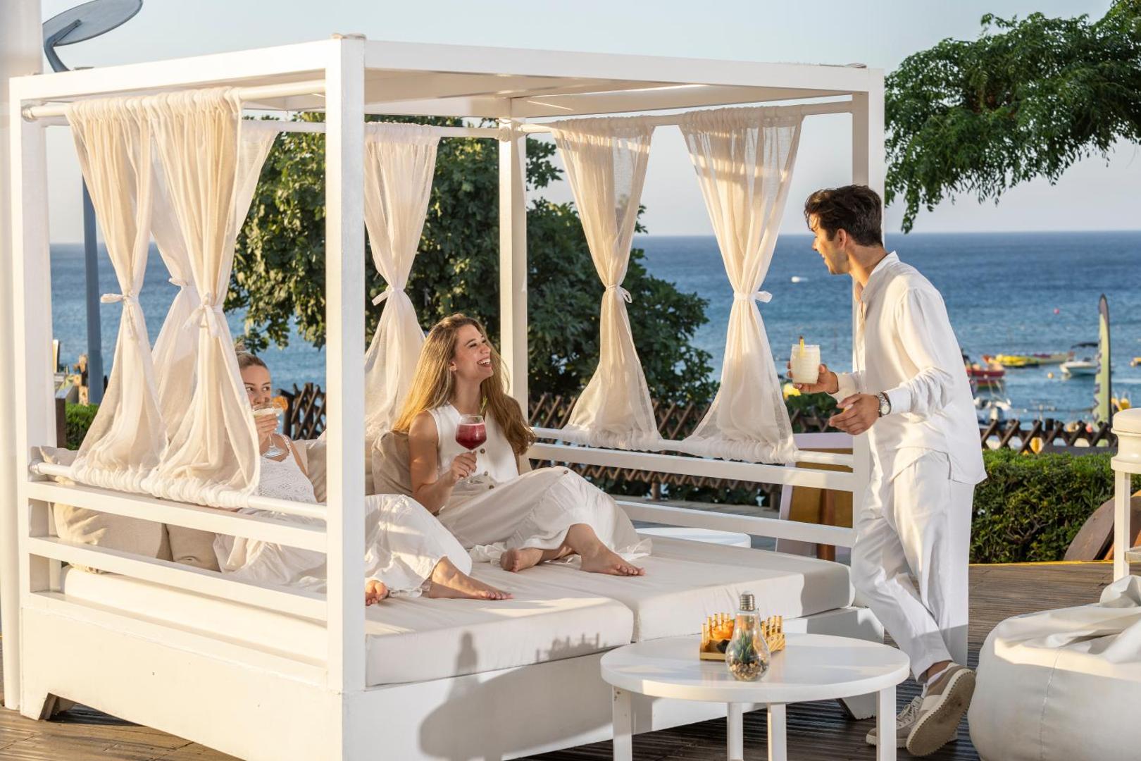 Capo Bay Beach Hotel atpūtas zona ar baltām nojumēm pie jūras Protarasā, Kiprā — viesi bauda kokteiļus uz mīkstiem zviļņiem