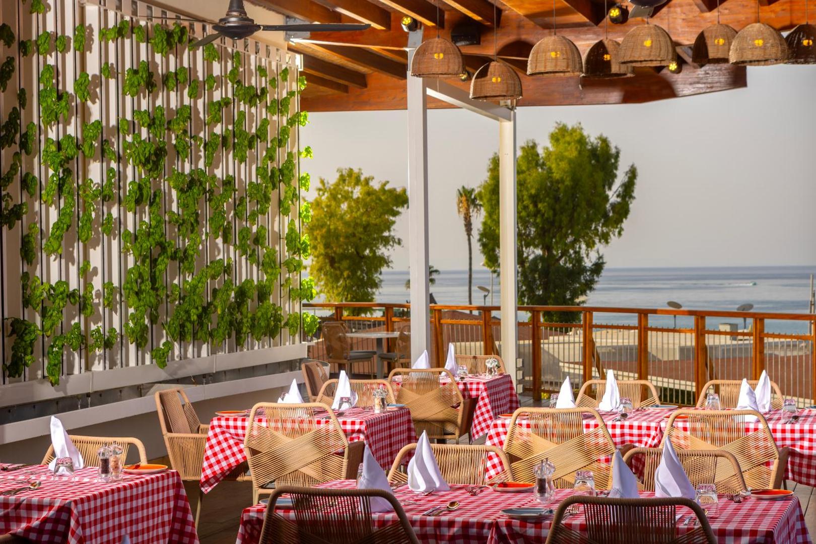 Capo Bay Beach Hotel itāļu stila restorāns ar sarkaniem rūtainiem galdautiem un skatu uz jūru, dekoratīva zaļumu siena