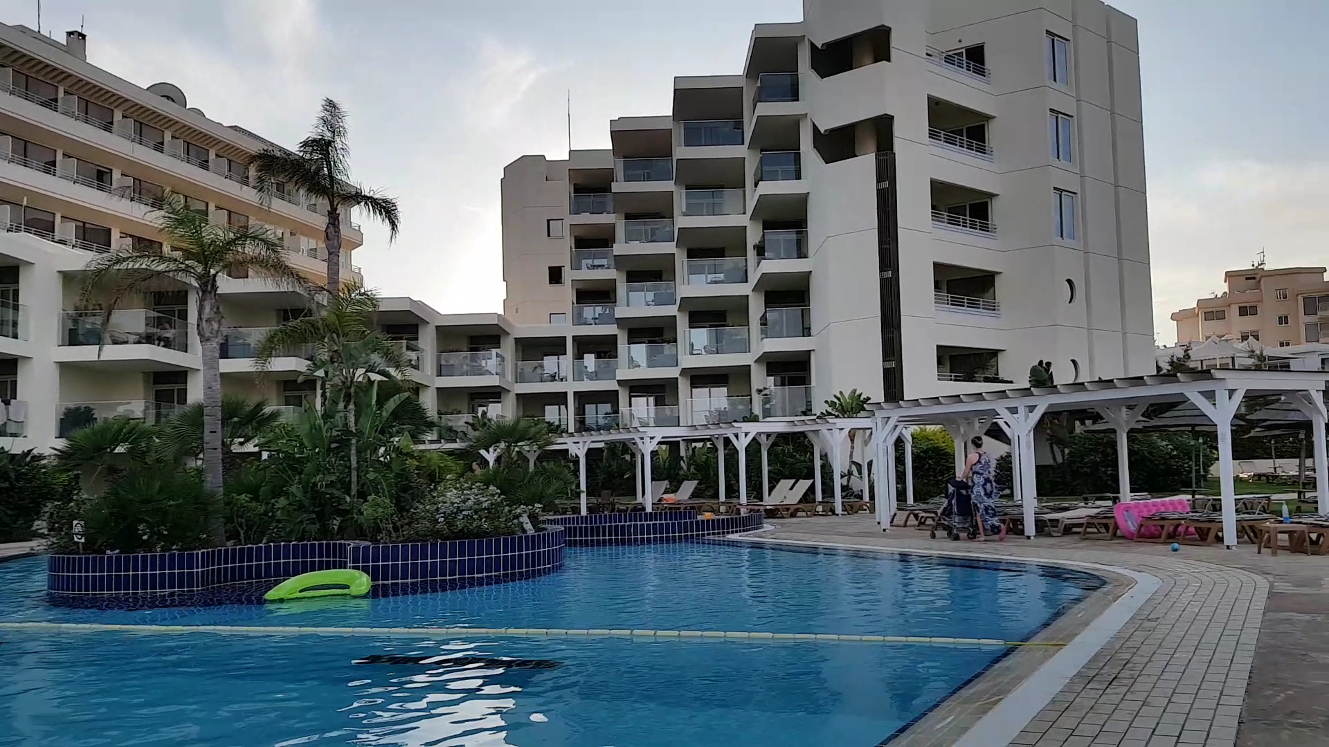 Capo Bay Beach Hotel baseina zona vakara gaismā — balti saulessargi, palmas un viesnīcas numuru balkoni Protarasas kūrortā