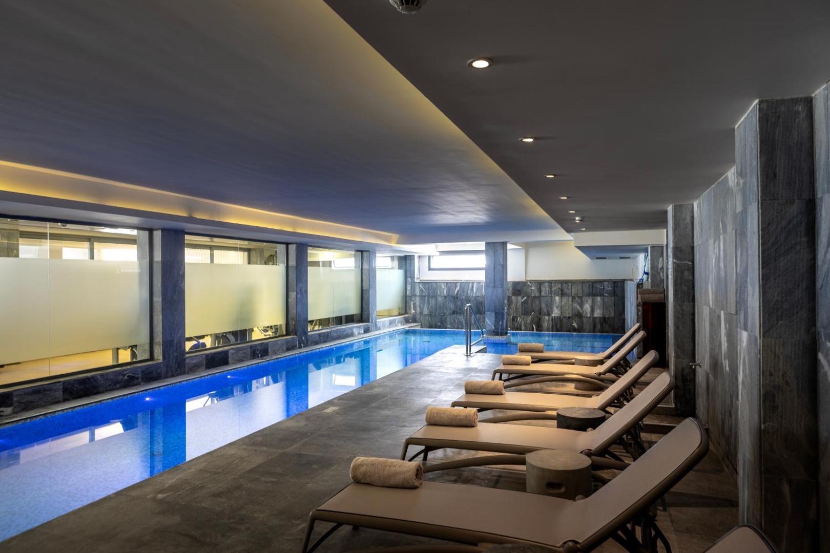 Capo Bay Beach Hotel iekštelpu baseins ar zilu apgaismojumu un ērtiem zviļņiem — moderna spa zona relaksācijai