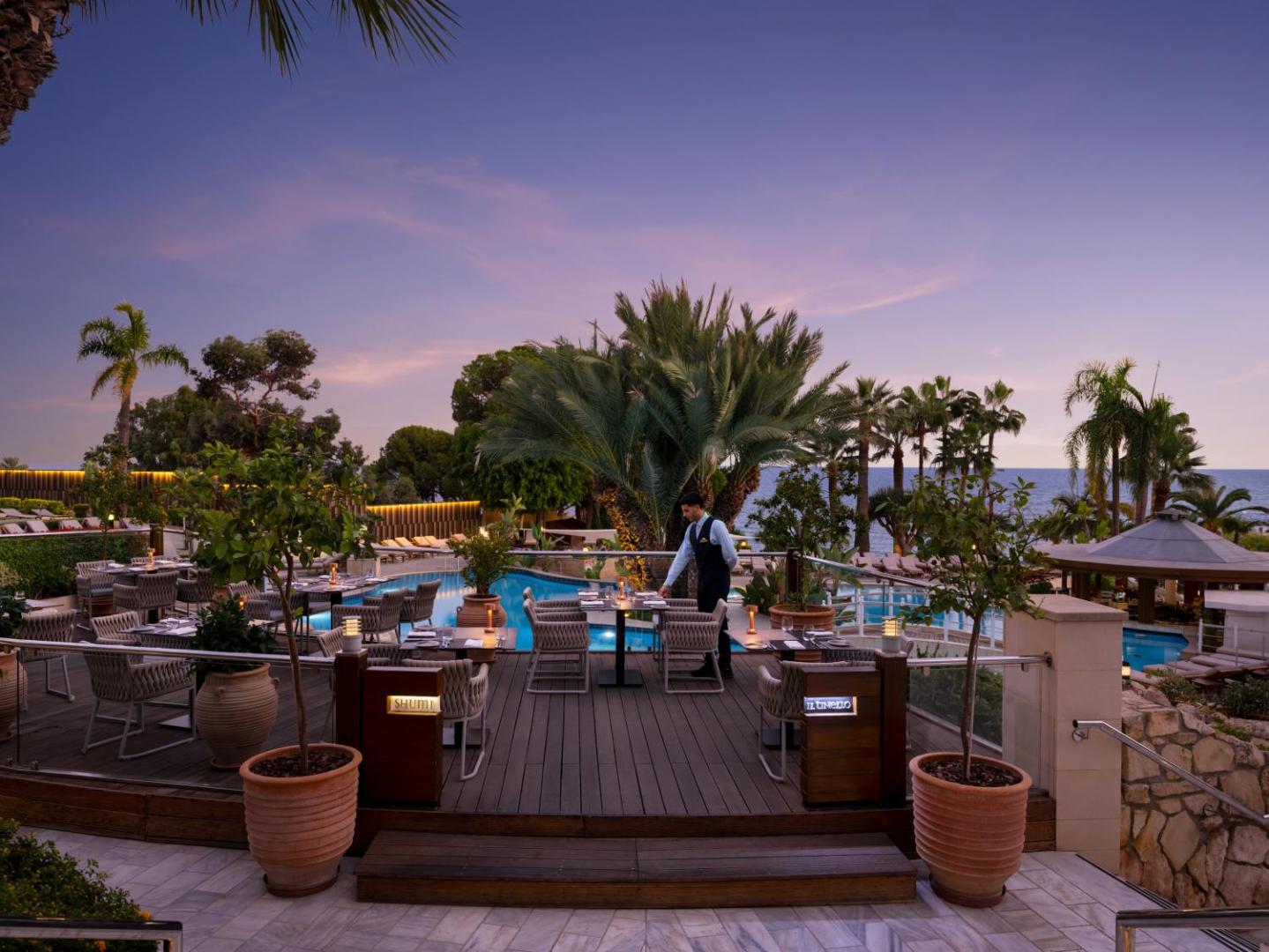 Mediterranean Beach Hotel āra restorāna zona pie baseina vakarā — koka klājs, palmas un skats uz piekrasti saulrietā