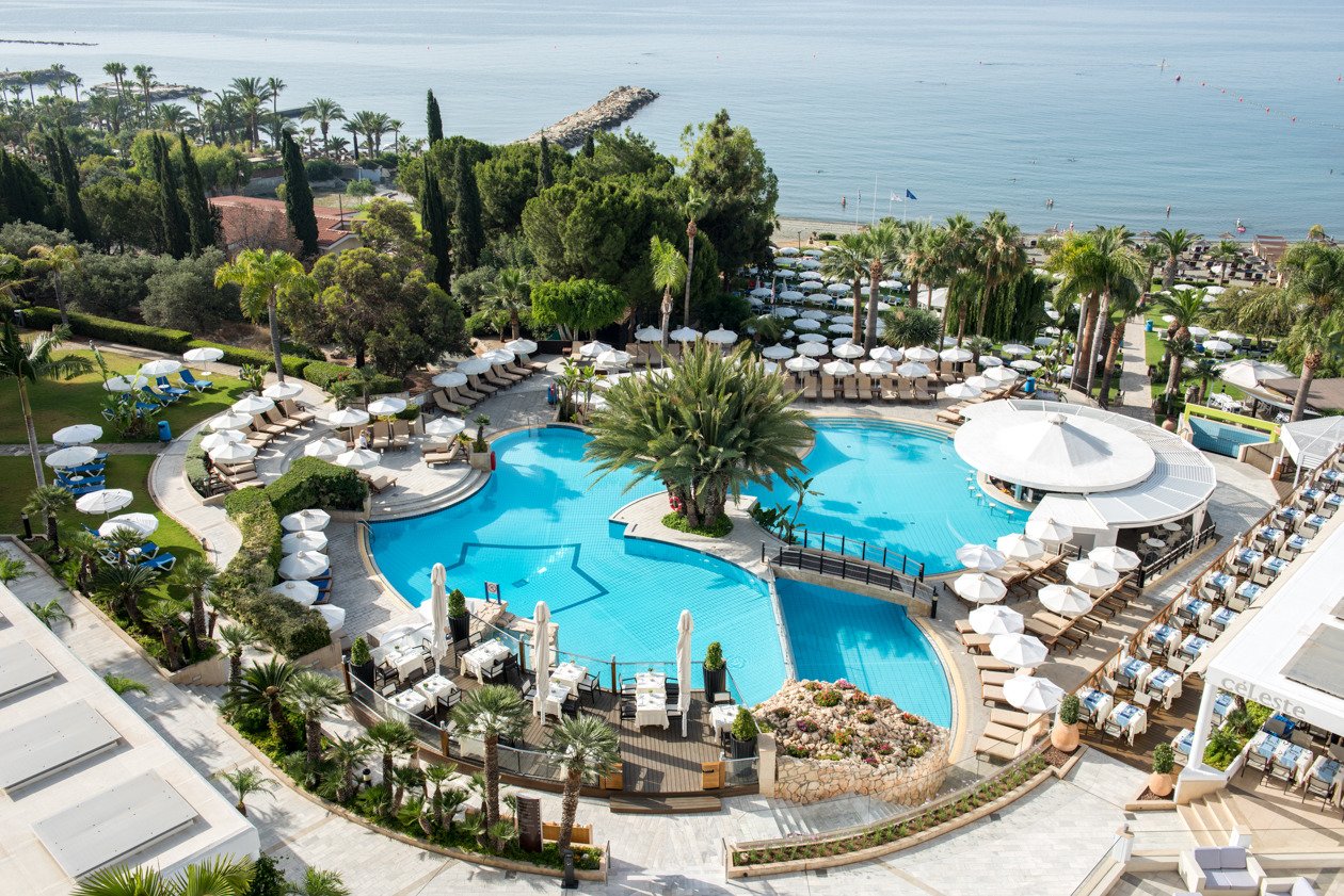 Mediterranean Beach Hotel āra baseinu komplekss Limasolā, Kiprā — skats no augšas uz baseiniem, palmām un Vidusjūras piekrasti saulainā dienā